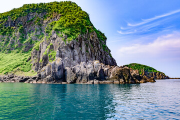北海道・世界自然遺産の知床半島を巡るクルーズ風景。6月、青空と薄い雲が入り混じる空の下、雄大な断崖や原生林、海が広がる景観。