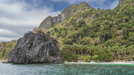 A picturesque sheer cliff in the turquoise ocean. Palm trees on a sandy beach. Green tropical vegetation on a mountainside. Boats on the shore. Philippines. Palawan. El Nido. Seven Commandos beach