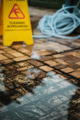 Folded caution wet floor cleaning in progress sign and a gardening hose reel on wet, flooded, mossy and dirty patio outdoors