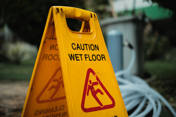 Two-sided caution wet floor cleaning in progress signboard, gardening hose reel and a garbage container on wet, flooded, mossy and dirty paving blocks outdoors