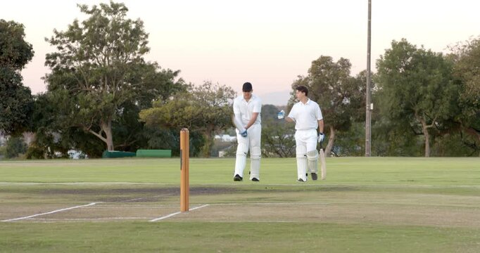 Emerging crease male teammates with bats adjusting gloves tapping stumps for break at dusk on pitch