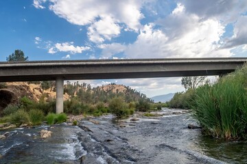 Serene river flowing under a bridge with lush greenery and a bright blue sky in the background