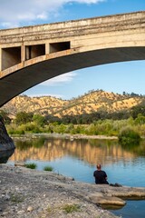 Man sitting by a serene river under a stone bridge, surrounded by lush greenery