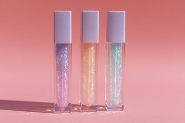 Three tubes of shimmering lip gloss in purple, peach, and blue shades arranged neatly on a pink background