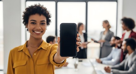 Happy African-American woman presents a mobile phone with a blank screen while colleagues meet in the background.