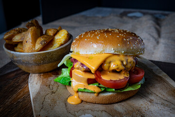 A hamburger with sesame seed bun, meat, melted cheese, lettuce, and slices of tomato is on a wooden board, along with potatoes.