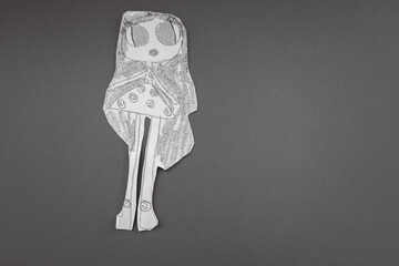 A black-and-white paper doll, cut out from a drawing, stands on a solid gray background. This...