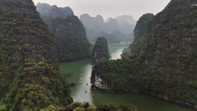 Traditional sampan boats navigate the winding Sao Khe River between towering limestone karsts in Trang An, Ninh Binh, Vietnam, captured by ascending drone shot.