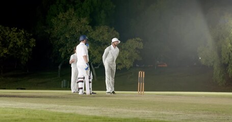 Diverse cricket players surveying batsman on floodlit pitch bowler running in bowling at stumps - Powered by Adobe