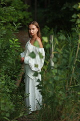 Naklejka premium Young beautiful woman in a long white cotton dress in the forest near the river behind the bushes