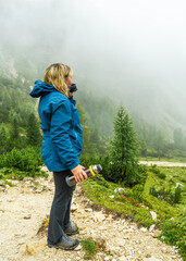 Side view of woman in rain jacket drinking hot tea while hiking in the foggy Dolomites. Rain and...