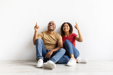 Happy African American couple sitting on floor pointing upward, smiling while looking at blank...