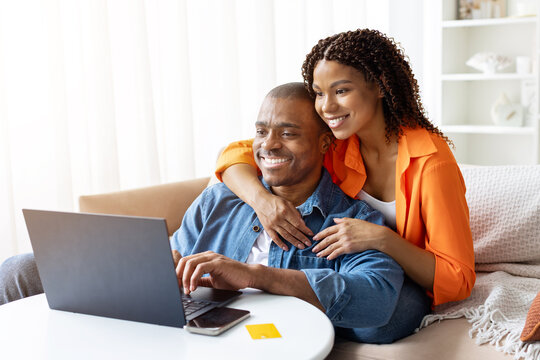 Smiling young African American couple using laptop at table at home, cheerful black woman hugging man from behind, both looking happy together