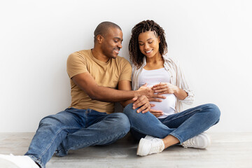 Smiling African American couple sitting on floor, man gently touching pregnant wifes belly, sharing...