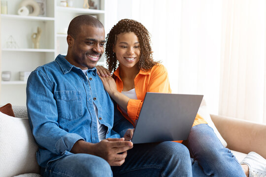 Smiling young African American couple relaxing at home on couch, browsing laptop together, enjoying time and sharing a joyful moment indoors, copy space