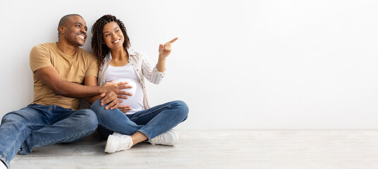 Smiling African American couple sitting on floor, man holding pregnant wifes belly while she points...