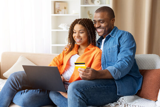 Happy young African American couple sitting on couch at home, online shopping with laptop and credit card, smiling together while booking vacation or paying bills
