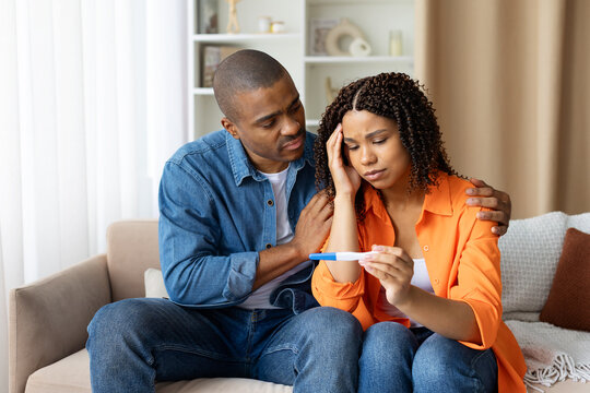 Worried African American couple sitting on couch, holding pregnancy test and looking upset during a tense moment at home, closeup shot