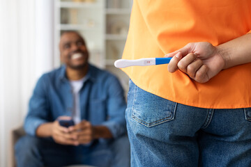 African American woman hiding positive pregnancy test behind her back, smiling while man in...