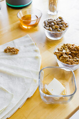 illustrating the process of making homemade baklava in a cozy kitchen environment. On a wooden table by the window, the ingredients are neatly arranged: bowls of walnuts, almonds and pistachios, a