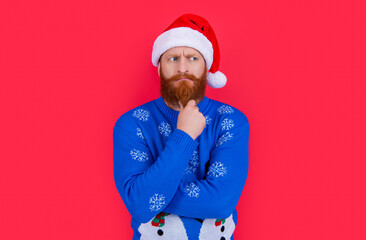 caucasian pondering man in new year sweater and santa hat isolated on red background.