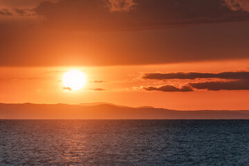 Golden sunset reflecting on calm sea with distant mountains and clouds creating a dramatic and...