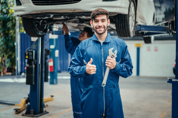 Mechanic in blue uniform smiling with wrench and thumbs up, colleague working in background, symbolizing teamwork, service quality, and professionalism in automotive repair.