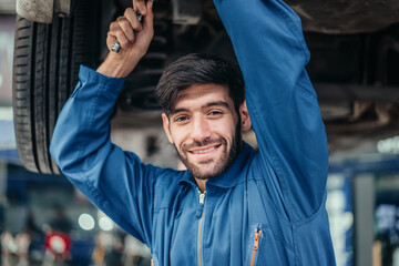 Male mechanic in blue uniform working under car with wrench, smiling at camera, representing trust, professional service, and customer satisfaction in automotive repair.