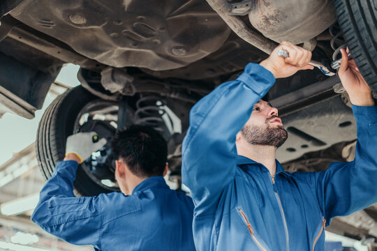 Two mechanics in blue uniforms repairing suspension and exhaust on lift, representing teamwork, quality service, and repair expertise in automotive industry. - Powered by Adobe