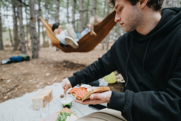 A young man is eating a sandwich during a picnic in a tranquil forest, while others relax nearby in...