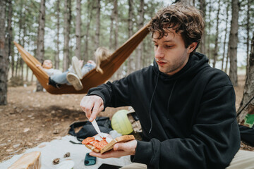 A man in casual attire spreads ingredients on bread at a picnic in the forest, with friends...