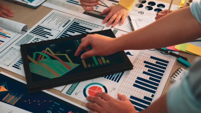 A diverse business team collaborates in a modern office, analyzing various graphs and charts on a tablet and printed reports, showcasing teamwork and strategy. SACTR