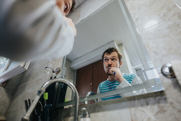A young man brushing his teeth in front of a bathroom mirror, reflecting his routine and hygiene...