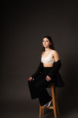 A girl in a pantsuit and a short top in a studio on a black background sits on a light wooden chair.