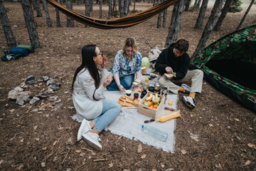 A group of three friends gathers for a picnic in a serene forest setting, surrounded by camping...