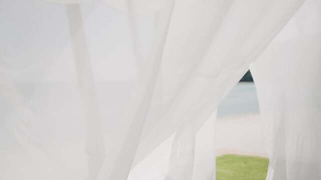 A view of the tranquil beach and ocean seen through flowing white sheer fabric. The soft, elegant drape creates a serene and romantic atmosphere, perfect for a wedding, special event, or relaxing tro