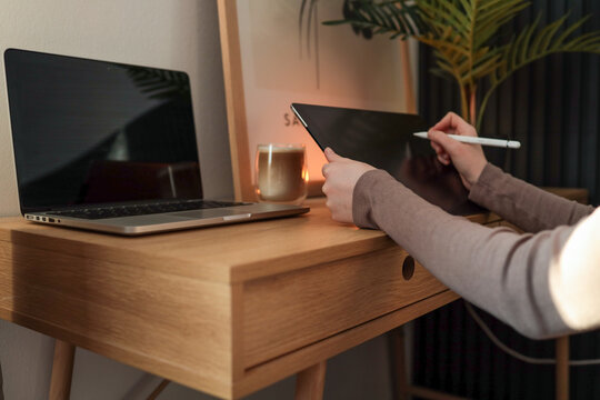 Side view of a person working at a wooden desk with a tablet and white stylus. A closed laptop, a glass cup of latte, and warm lighting create a cozy and modern workspace atmosphere