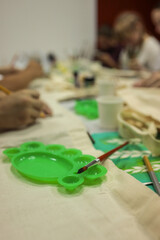 Close-up of art workshop with paintbrushes, green plastic palettes, and people painting on fabric bags at a shared table, creative group activity
