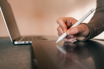 Close-up of hand holding stylus and working on digital tablet with laptop in background on desk