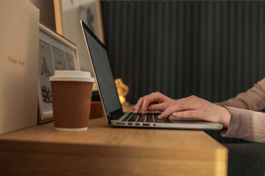 Person typing on laptop at wooden desk with takeaway coffee cup, framed artwork, and warm ambient lighting in cozy workspace