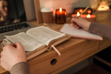 Open Bible on wooden desk with a person writing in a planner using a gold pen, surrounded by lit candles, a coffee cup, and a laptop, creating a warm and peaceful study atmosphere