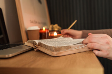 Open Bible on wooden desk with a person writing in a planner using a gold pen, surrounded by lit candles, a coffee cup, and a laptop, creating a warm and peaceful study atmosphere