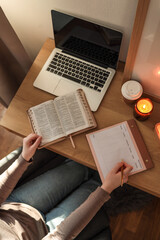 Person sitting at a wooden desk with an open Bible, a laptop, and a weekly planner, writing with a...