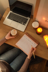 Top view of a person sitting at a wooden desk with an open laptop, holding a glass of coffee in one...