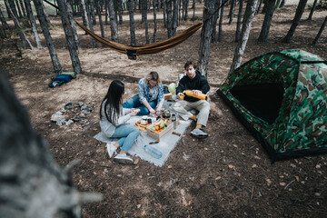 A group of friends enjoying a camping trip, surrounded by nature, sharing food and laughter near a tent and a hammock set up in a forest clearing.