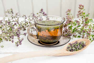 Homemade Oregano, Origanum vulgare herbal tea in a tea cup. Steaming hot drink with dry and fresh Oregano flower blossoms around, white wood board minimal background.