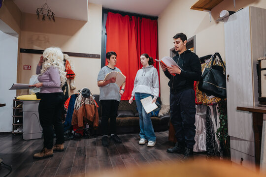 Actors engaged in a rehearsal backstage, holding scripts and preparing for a theatrical performance. A casual setting with costumes and props enhances the preparatory atmosphere.
