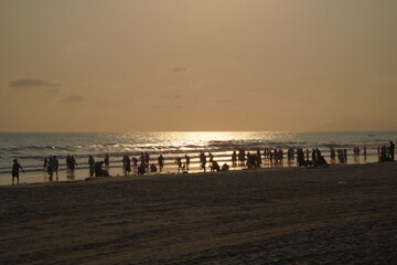 A beach scene at sunset, with people silhouetted against the golden glow of the setting sun over the sea