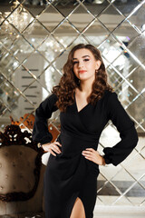 A woman in an elegant black dress stands indoors next to a luxurious chair and a mirrored wall. Her...