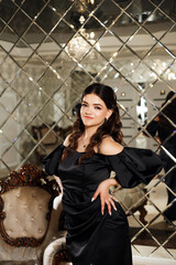 A woman standing in an elegant setting. She is wearing a black satin dress with puffy sleeves and a...